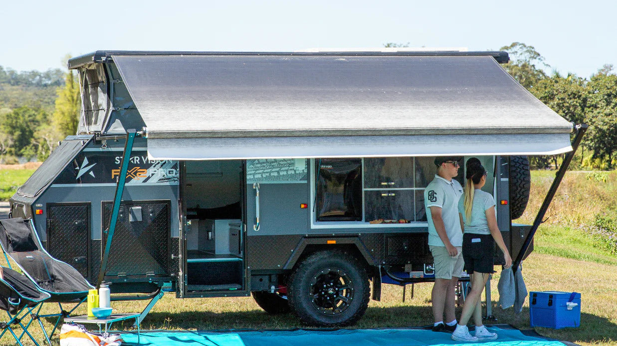 A couple stands beside a camper trailer, smiling and enjoying their outdoor adventure together.