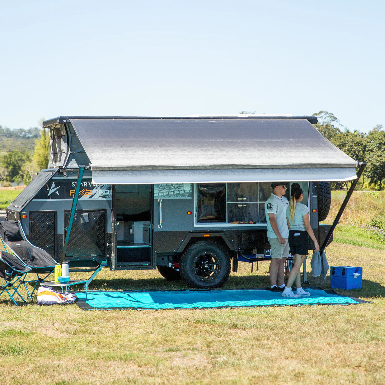 A couple stands beside a camper trailer, smiling and enjoying their outdoor adventure together.